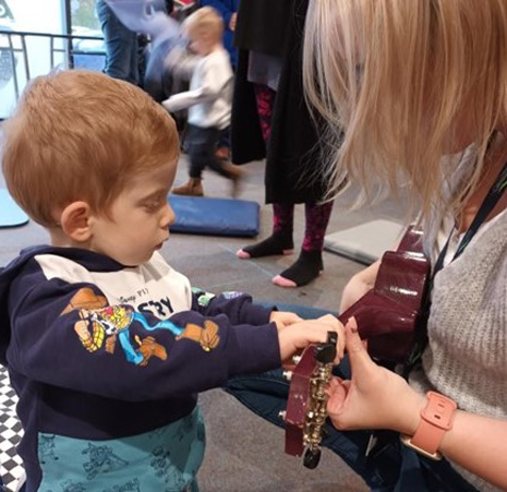 A young boy with ginger hair playing a ukulele offered by a woman with blonde hair