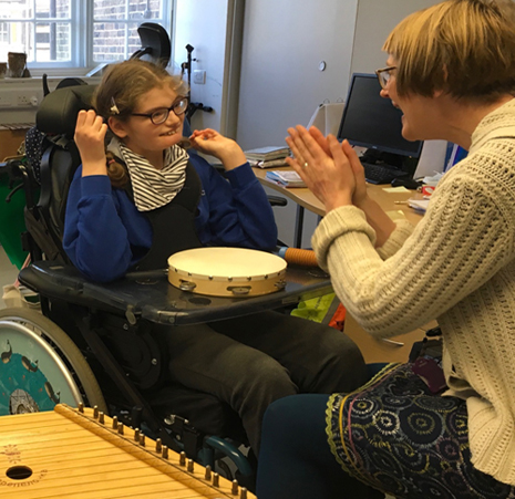 A teenage girl in a wheelchair with a small drum in front of hair, opposite a woman with short hair clapping
