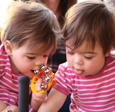 two small girls with short brown hair wearing matching pink stripy shirts, one holding and chewing on a small bell toy with an orange handle and silver bells.