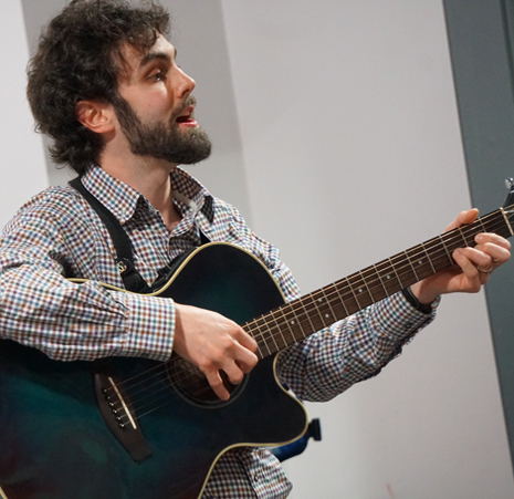 A man with curly dark hair and a beard wearing a blue check shirt playing a dark green guitar