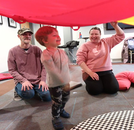 A small boy smiling under a red parachute, with a man in a cap and a woman in a pink jumper smiling behind him